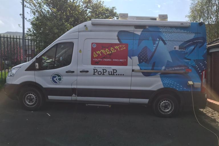 Picture of the Streetz Detached bus, a white and blue van with a red sign with the project's name, parked in a parking lot in front of a yard.