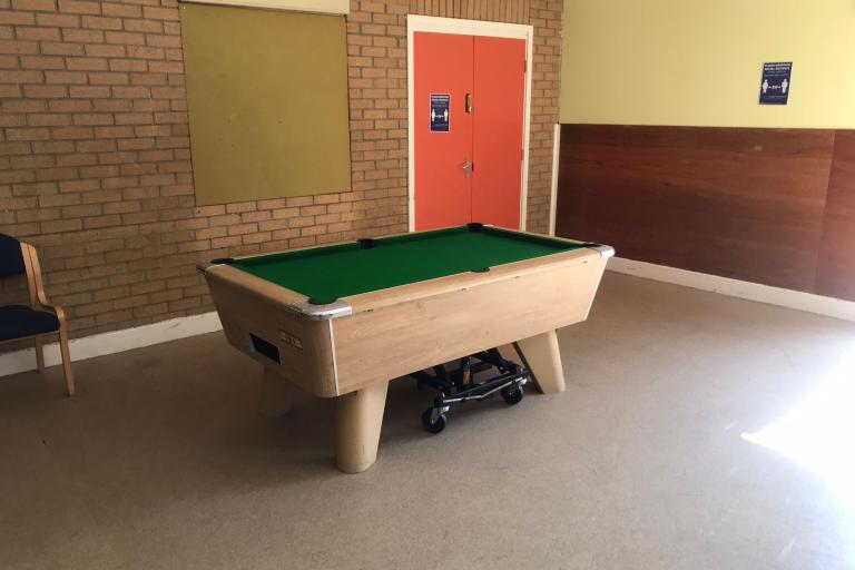 One of the youth rooms in the California Youth Centre. A pool table stands in the middle of the picture, a chair in the left corner and an exit double door in the right far corner.