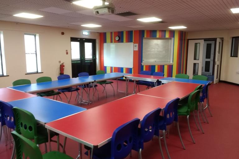 Spacious room with a large conference table for twenty people taking most of the space. The table is blue and red, the chairs are of different colours and the far back wall is painted in multiple colours as well.