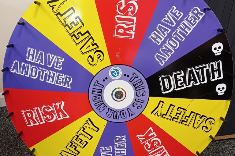 The drug wheel used in one of the activities of the Youth Work Plus. It is a spinning wheel which  has the words risk, safety, have another and death, placed on different tiles in an alternating manner