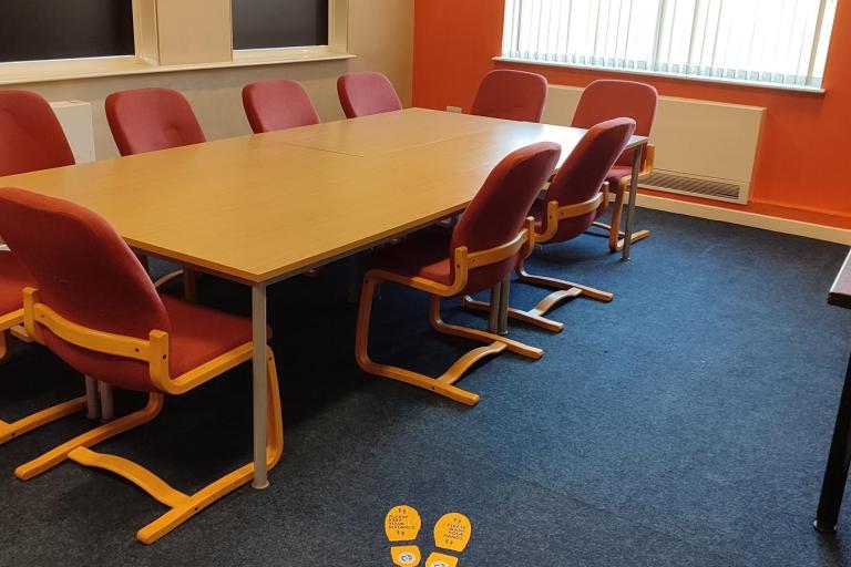 Conference room at the Grangetown Youth Centre. In the centre of the room there is a large table for twelve people, surrounded by chairs. Next to the right hand side wall, there are two desks with computers.
