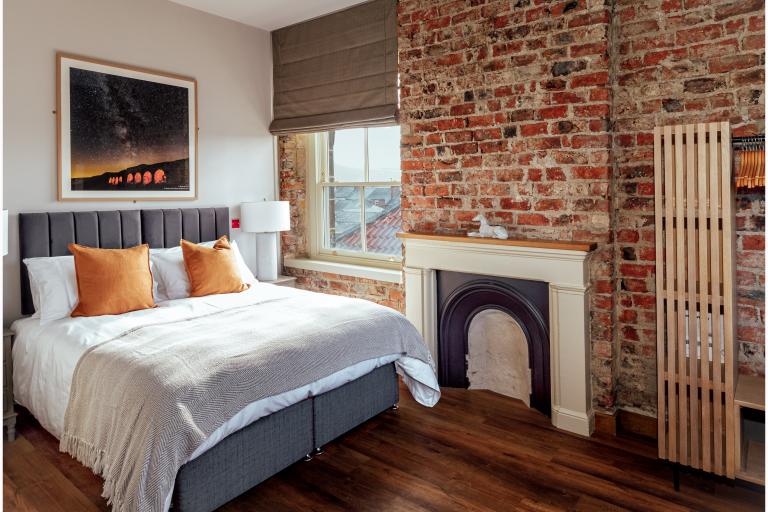 A photograph of a bed next to a window and exposed brick wall. 