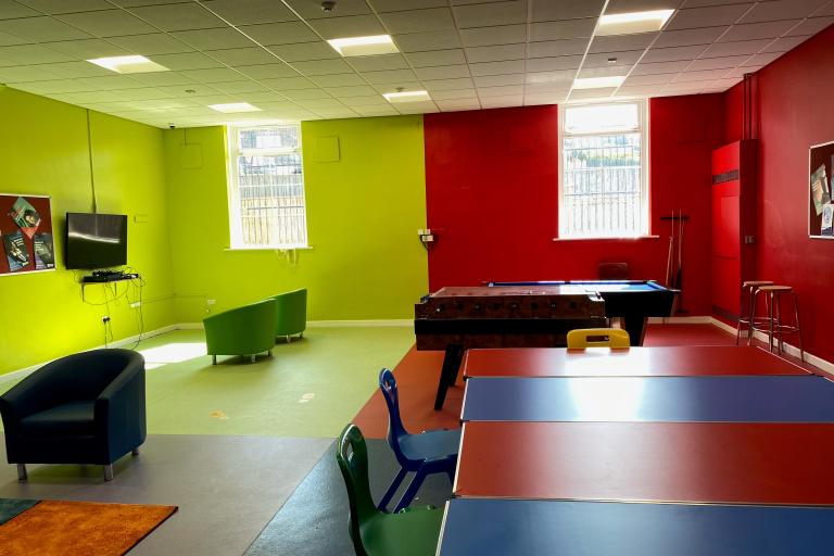 Same youth room from the Lingdale Youth with the green and red sections visible. In the red section, on the back right hand side, there is a foosball table and a pool table. In the green section, there are two green chairs in front a TV. There is a window placed in each of the two sections of the room. 