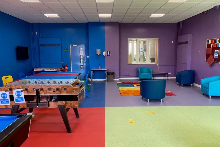 Youth room In Lingdale Youth Centre. A room divided in four sections painted in four different colours: blue, purple, red and green.  In the blue section, at the back left hand side there is an exit door, four rectangular red and blue tables put together with four chairs on each side. In the purple section, back right hand side, there are four armchairs of different shades of blue, a window towards the hallway, and a board with pictures and drawings on it. In the foreground, there is s foosball table.