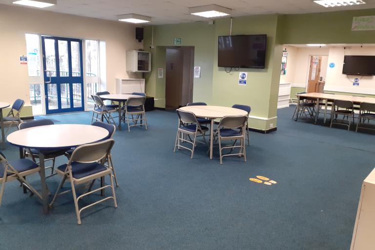 Lounge area in Loftus Youth Centre. Three round tables surrounded by chairs in the foreground in front of a TV. A second room in the background with a long table surrounded by chairs standing in front of a second TV.