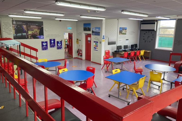 Main room of the centre. A picture taken from a balcony with several round blue tables surrounded by yellow and red chairs placed across a large room, three computers placed at the far left corner, two couches in the foreground, posters and inspiring quotes on the walls.