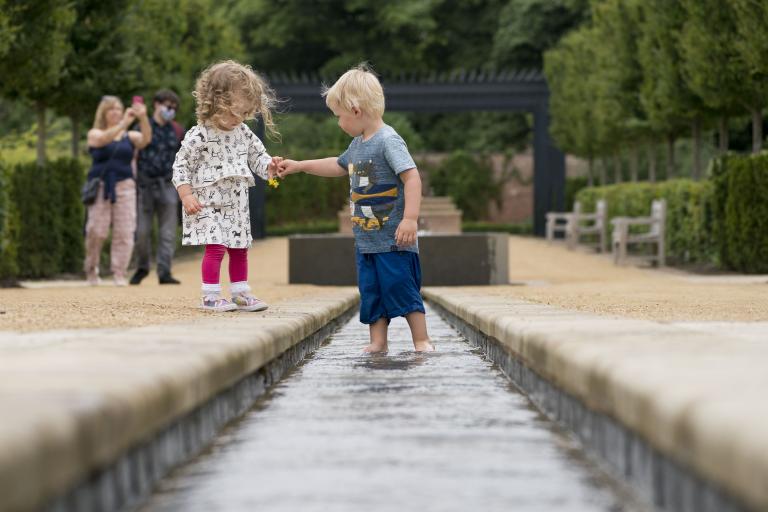 A little boy holding the hand of a little girl while playing in the water feature