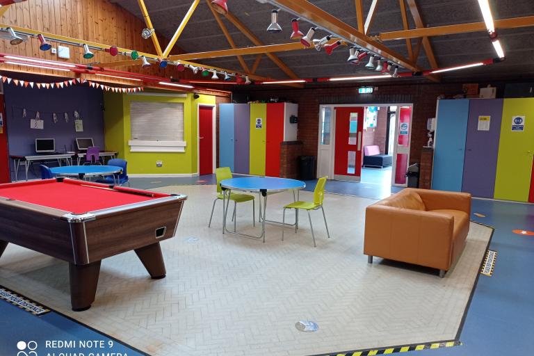 Image of the play room from the centre with a pool table, a couch, two computers in the back and round tables with chairs and wardrobes along the walls 