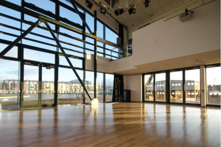 Picture of the main hall in Tuned In, a spacious room with floor to ceiling windows on one of the walls, wooden floor and spotlights on the ceiling.