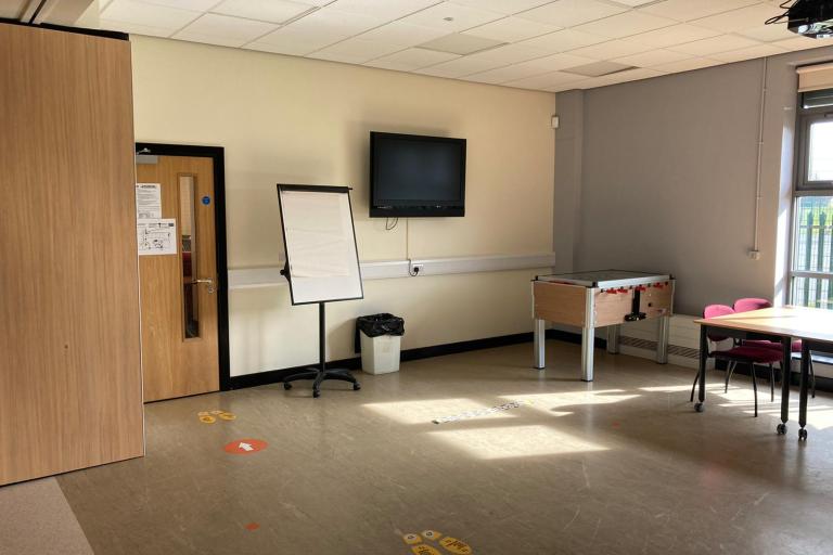 Picture with the training room at 25K Centre. On the left hand side there is a TV and a flip chart easel. In the back there is a small foosball table and two desks with chairs.