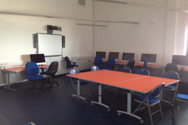 ICT room, there is a large table for eight in the middle of the room, along the walls there are desks with computers and on the wall on the left hand side there is a projector screen.