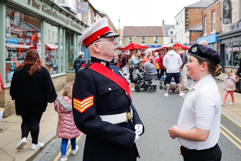 Man in military uniform talking to a young boy at the Chaloner Street Market. In the background there are crowds of people and market stalls.