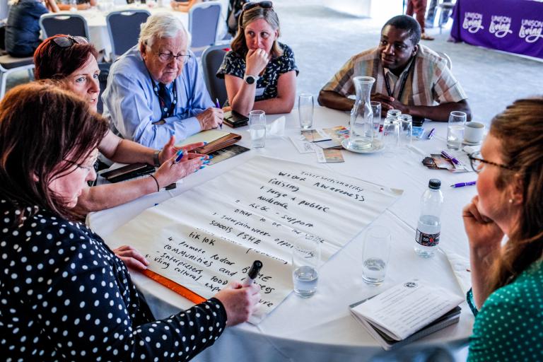 A group of people gathered around a table talking to each other and taking notes on a large piece of paper.
