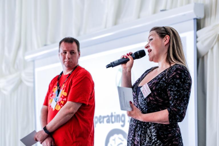 A woman dressed in a black dress and a man dressed in a red t-shirt standing on a stage with a powerpoint presentation behind them. The woman is talking to a mic.