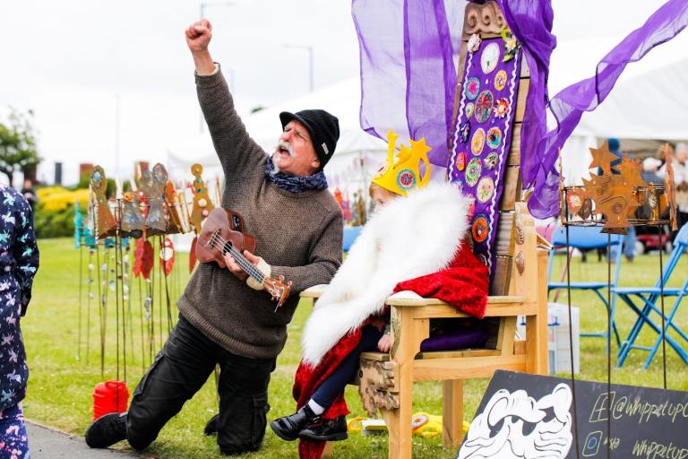 A man playing the ukulele next to a child dressed in royal clothes and sitting on a throne.