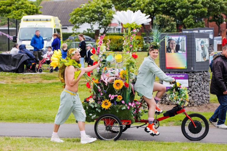 Two performers, a man and a woman, at Zetland Park. The woman is riding a bicycle decorated with flowers while the man walks next to her caring a giant bouquet of flowers.