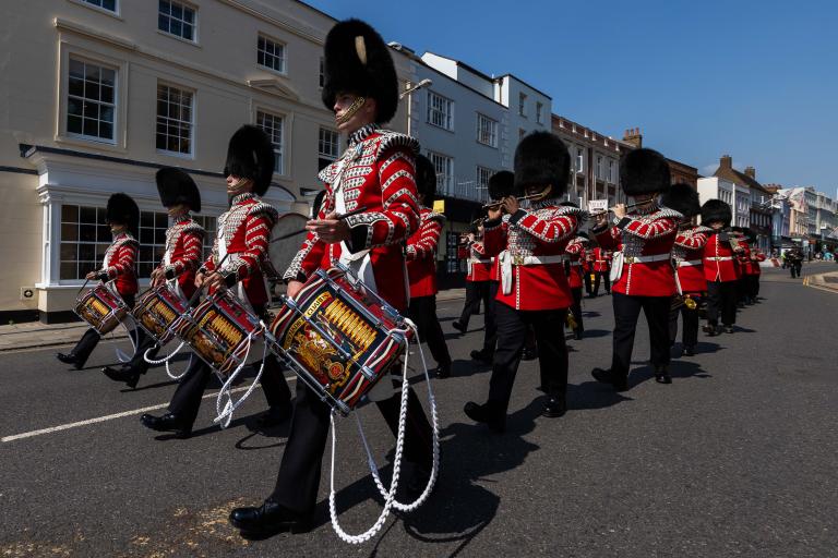 Grenadier Guards dressed in ceremonial uniforms marching on the streets. The front row is playing military drums while the following rows are playing the flutes.  