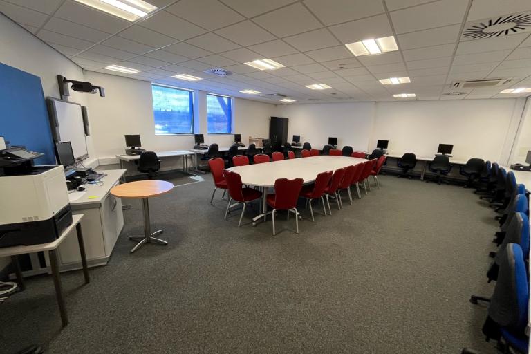 Image of the ICT Suite at Tuned In, a lare room with a board table surrounded by chairs in the middles and desks with computers around the walls. At one end of the room there is a large screen and projector