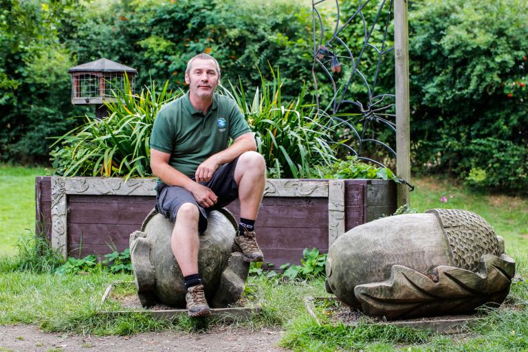 Image of park ranger Paul Murphy wearing a green polo shirt. Paul is sat on a wooden sculpture.