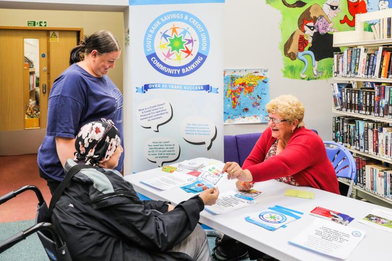 Image of a lady dressed in a red shirt giving advice to two residents at the cost of living drop-in session at Grangetown library.