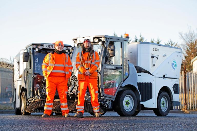 Image of two Streetscene officers dressed in orange overalls with two of the new street sweepers.