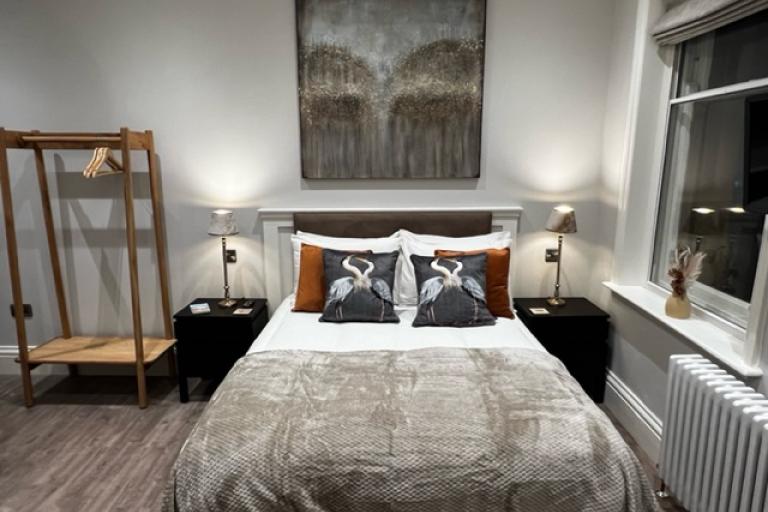 Image shows one of the accommodation bedrooms for visitors in the Barclays Bank in Loftus. Inside the room there is a double bed with nightstands on both sides, a painting hanged above the bed and a clothes rail.