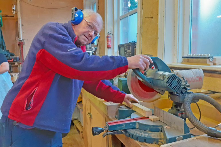 image of a volunteer dressed in a blue and red jacket, cutting wood for a bird box
