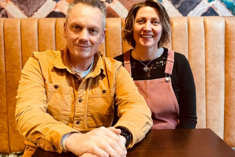 Image of a man wearing a yellow shirt and a woman wearing a black shirt and red apron sat at a table.