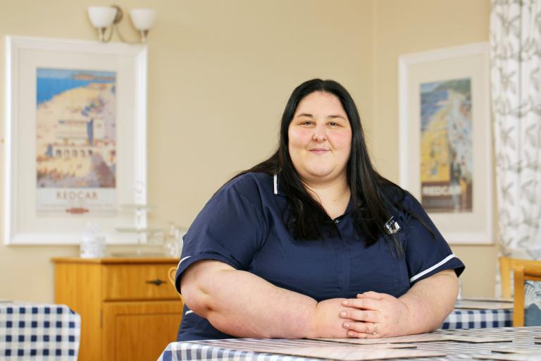 Picture of a woman wearing a blue nurse uniform, sat at a desk and smiling.