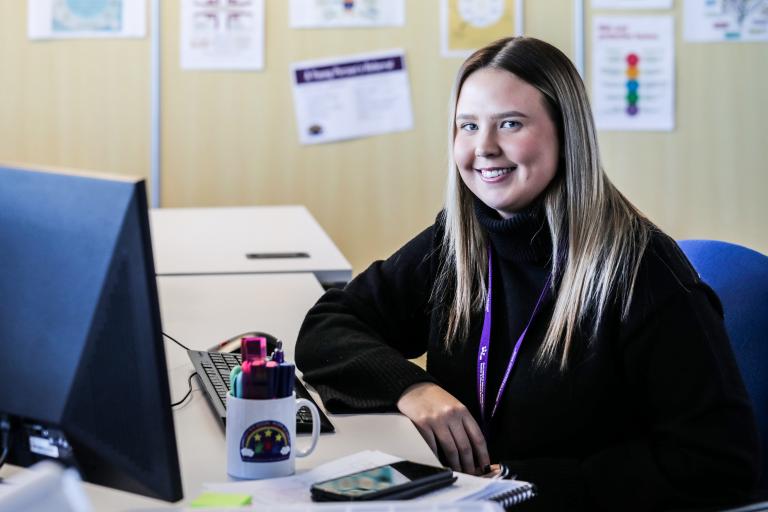 Image of a young woman sat on a desk and smiling at the camera.