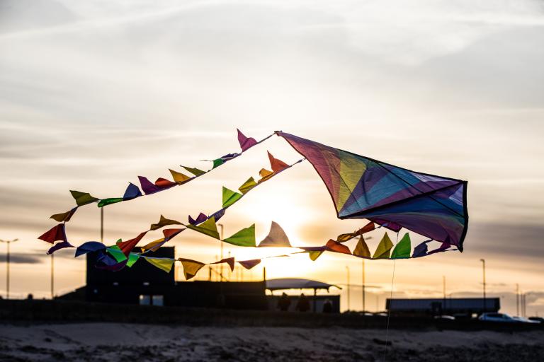 Image of a rainbow coloured kite flying in the sky with the sunset in the background.