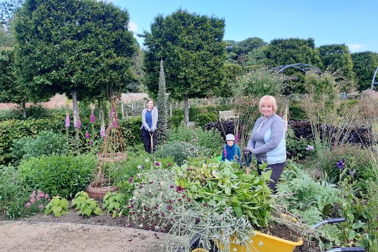 Image of three women tending to a garden. One is holding a gardening tool. There is a yellow wheelbarrow in the foreground.