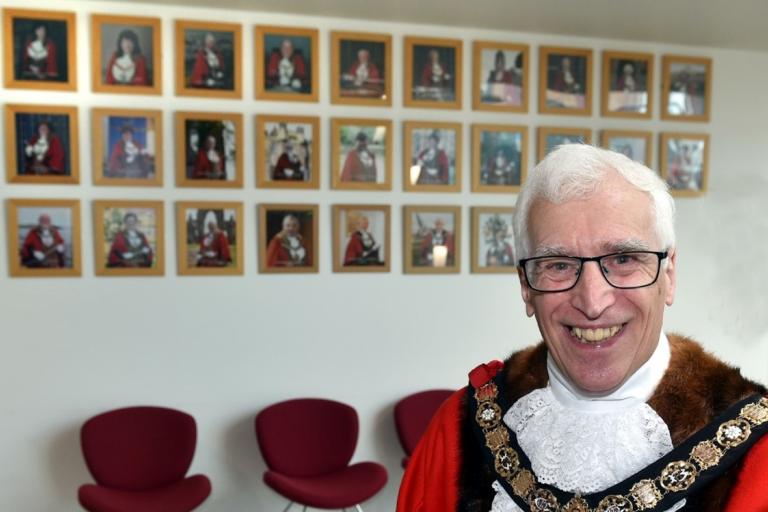Image of Cllr Ian Hart. Ian is wearing red mayoral robes and ceremonial chains. Images of previous mayors are in the background.