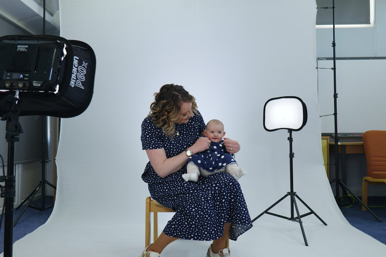 A woman in a blue dress sitting on a chair and holding a baby dressed in the same colour. Behind her there is a white background and studio lights.