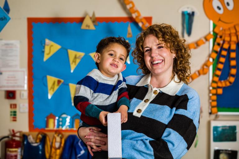 A woman holding her toddler in her arm, they are both looking and smiling at the camera. 