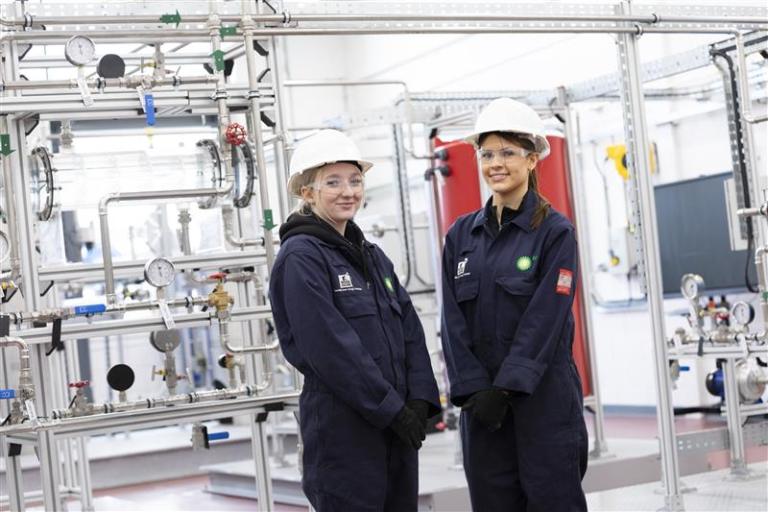 Image shows two young women wearing blue overalls and white helmets stood in front of pipework