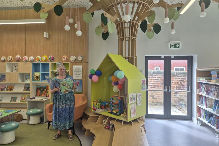 A woman holding a book standing in the middle of a play area from a library, with a decorative tree and bookshelves behind her.