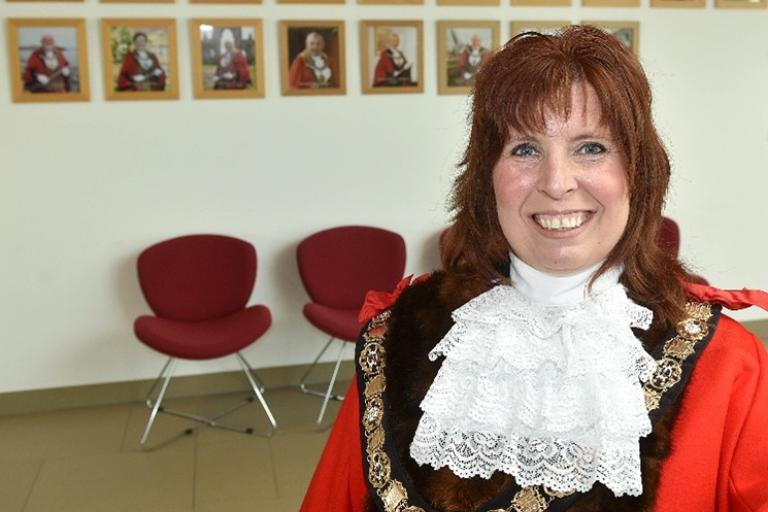 Image shows Cllr Cawley wearing her read Mayoral Robe standing in front of a wall with images of previous mayors