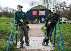 Image of two men wearing outdoor maintenance clothing. They are leaning on a blue bridge. 