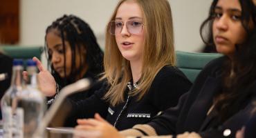 A young girl with blond hair and wearing glasses and a black shirt, sitting by a desk and talking