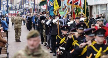 Image of an armed forces parade. A man in camo uniform is in the foreground with marchers in blue outfits in the background with various flags waving.