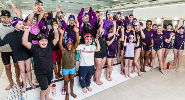 A group of children dressed in purple swim gear standing in front of an indoor pool with their hands raise. In the middle of the group stands the Paralympic swimming champion Maisie Summers-Newton MBE wearing the Great Britain Team t-shirt