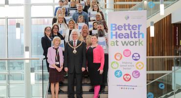 Image of a group of people stood together on some stairs. A sign to the left of the group reads 'better health at work'