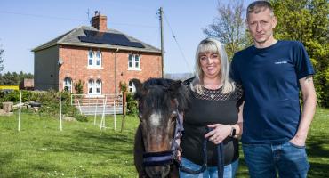 The picture shows a pony, a woman and a man standing in a green field with a red-bricked house and the blue sky in the background