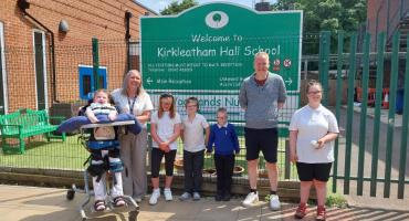 Two adults and five children, one of them in an accessible motorised wheelchair, standing in front of a sign reading Kirkleatham Hall School