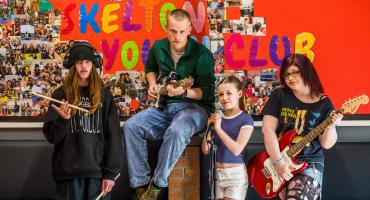 Two young men and two young women standing in front of a colourful mural, each one holding different instruments such as: drum sticks, a guitar, a mic and another guitar
