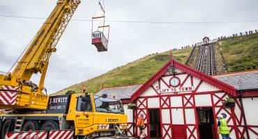A tramway carriage being lifted by a crane over a building towards two tramway rails that go up a hill 