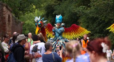 A performer dressed in a colour costume with wings that makes her look like she is riding a fictional giant bird is surrounded by crowds of people.