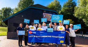 A group of people standing in front of a wodden building in a countryside park, holding a banner and multiple signs saying stop smoking