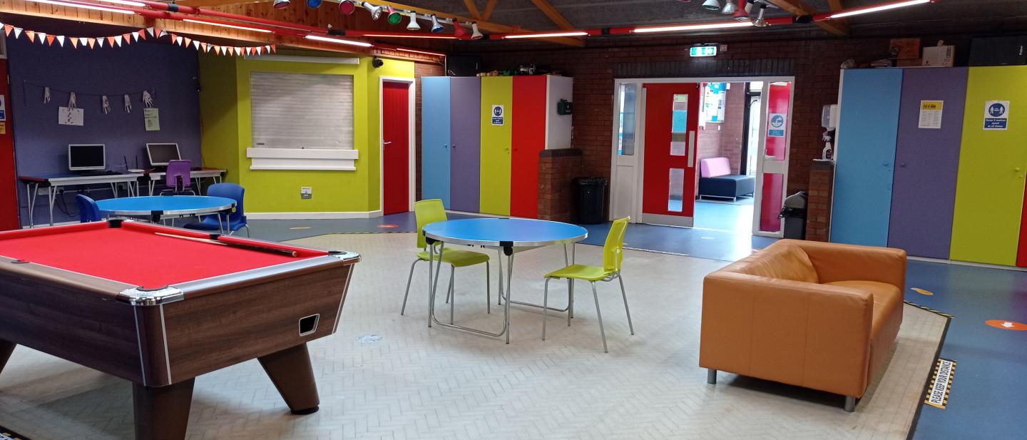 Image of the play room from the centre with a pool table, a couch, two computers in the back and round tables with chairs and wardrobes along the walls